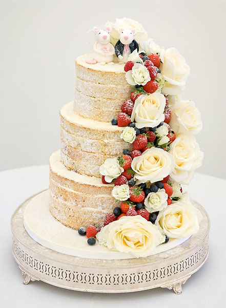 Three Tier Naked Wedding Cake with Layers of Vanilla Buttercream & Strawberry Preserve, with a Cascade of Large Fresh Ivory Roses & Berries. Topped with Bride & Groom Sugar Mice.