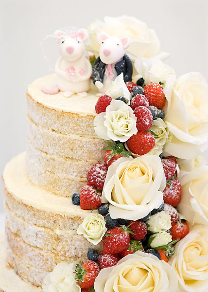 Three Tier Naked Wedding Cake with Layers of Vanilla Buttercream & Strawberry Preserve, with a Cascade of Large Fresh Ivory Roses & Berries. Topped with Bride & Groom Sugar Mice.